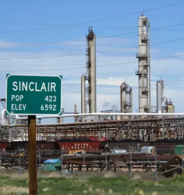 A green sign sitting in front of an oil refinery.
